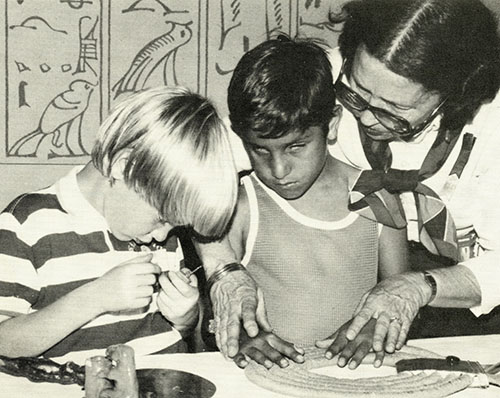 Two boys with visual impairments, one white and one brown, sit at a table that holds a few objects. A white woman stands behind one of the boys, guiding his hands around a circular object. The other boy holds a small object close to his face.