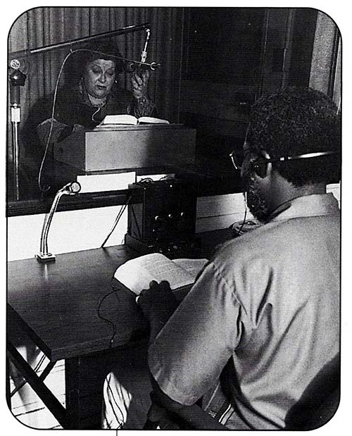 Argentinian narrator, Graciela Lecube sits in a recording studio, behind a glass window, in front of a microphone. A book is open in front of her, propped on a stand. In the foreground, facing Lecube, a Black man sits a desk with a book open. The man wears headphones.