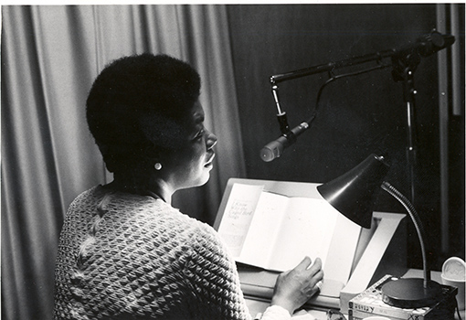 Maya Angelou sits in profile in front of a microphone and table lamp. Her book 