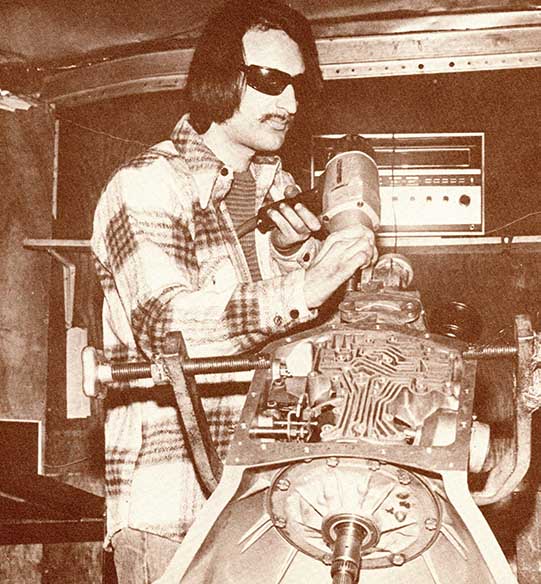 A white Vietnam War veteran who is blind uses a drill on a large metal structure that dominates the foreground of the image. A large radio is visible on a shelf behind him.