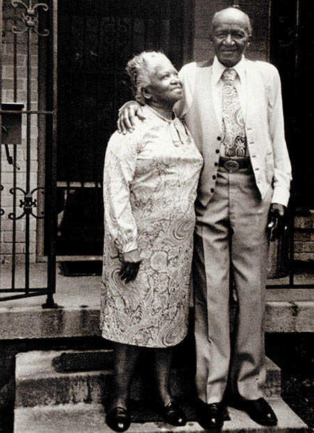 An elderly Black couple stand on the stoop outside their home. The couple are smiling. The wife looks up toward her husband, who has his arm around her shoulder.