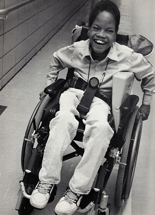 A smiling, young Black child with a visual impairment moves his wheelchair along a hallway.