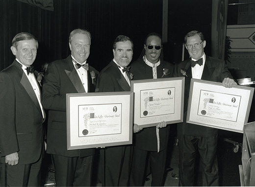 Stevie Wonder, smiling and dressed in formal attire, stands with four white men. The group holds three awards between them.