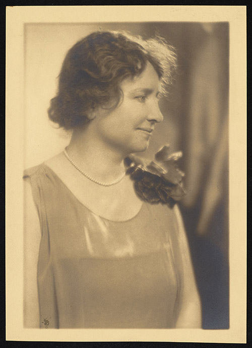 Sepia photograph of a half-length portrait of Helen Keller. Keller smiles gently, her face in profile. She wears a sleeveless boat-neck dress with a flower decoration on her left shoulder and a pearl necklace.