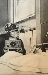 A white child who is blind lies in bed. Helen Keller sits on the far side of the bed, smiling and holding a radio set.