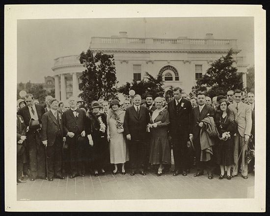 A cluster of over 30 delegates in formal attire stand outside the White House. Most of the visible delegates are white. They stand close together, with serious expressions on their faces.