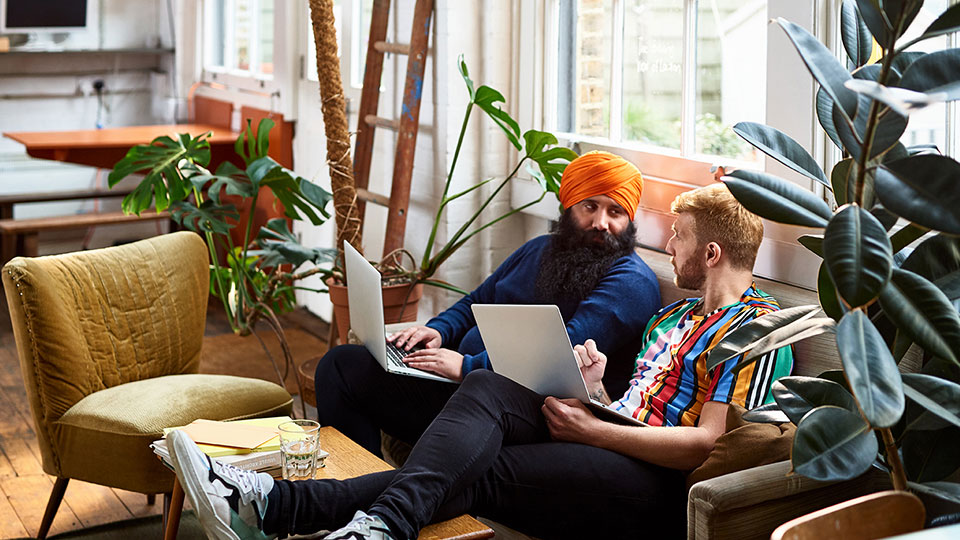 a pair of coworkers sitting on a couch holding laptops and talking