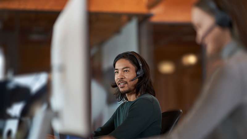 Pria dengan rambut panjang mengenakan headset, bekerja di komputer di kantor, dengan kolega di latar belakang