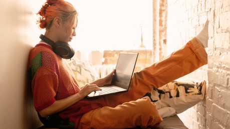 Een jonge vrouw met oranje haar zit comfortabel in de buurt van een raam met een over-the-ear hoofdtelefoon om haar nek en kijkt naar haar Windows 11-laptop.